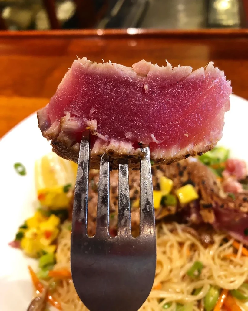 Rare seared tuna on a fork, with a blurred Asian-inspired meal behind.
