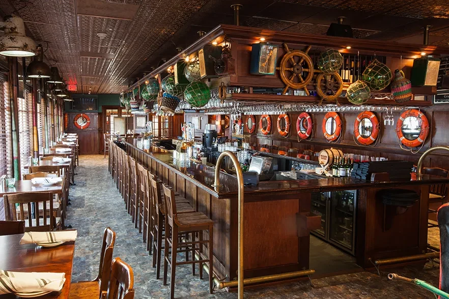 Empty nautical-themed restaurant interior with long bar and set tables.