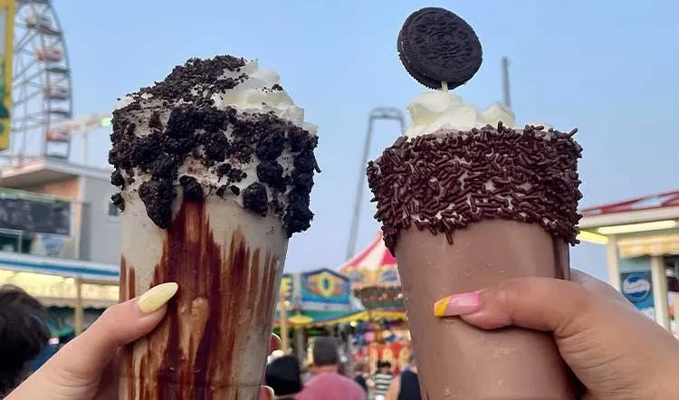 Two elaborately decorated milkshakes at an amusement park with a Ferris wheel.
