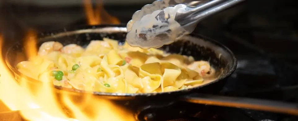 Tongs stir creamy pasta with peas and shrimp flaming in a dark pan.