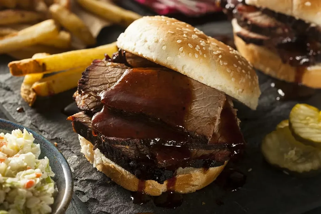 Sliced barbecue brisket sandwich with coleslaw and fries on a dark surface.