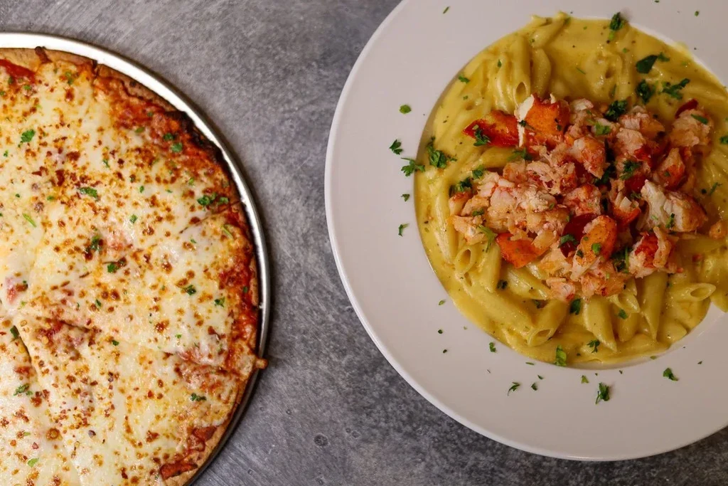Cheese pizza and lobster penne pasta side-by-side on a dark surface.