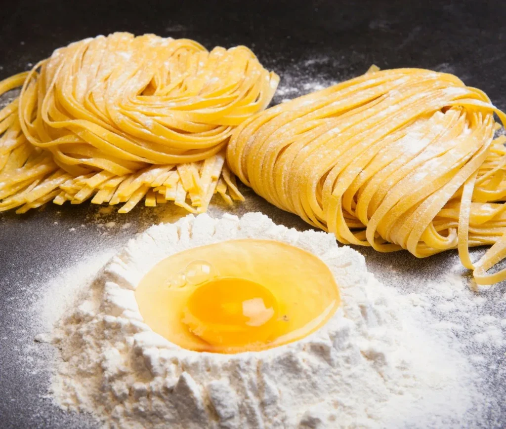 Fresh pasta nests, flour, and a cracked egg on a dark surface.