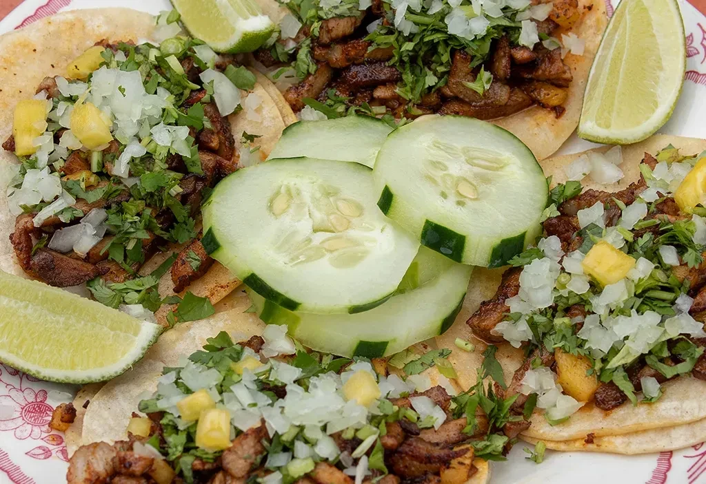 Close-up of al pastor tacos with onion, cilantro, pineapple, lime, cucumber.