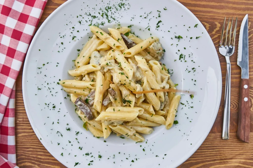 Creamy penne pasta with herbs, fork, knife on a wooden table.