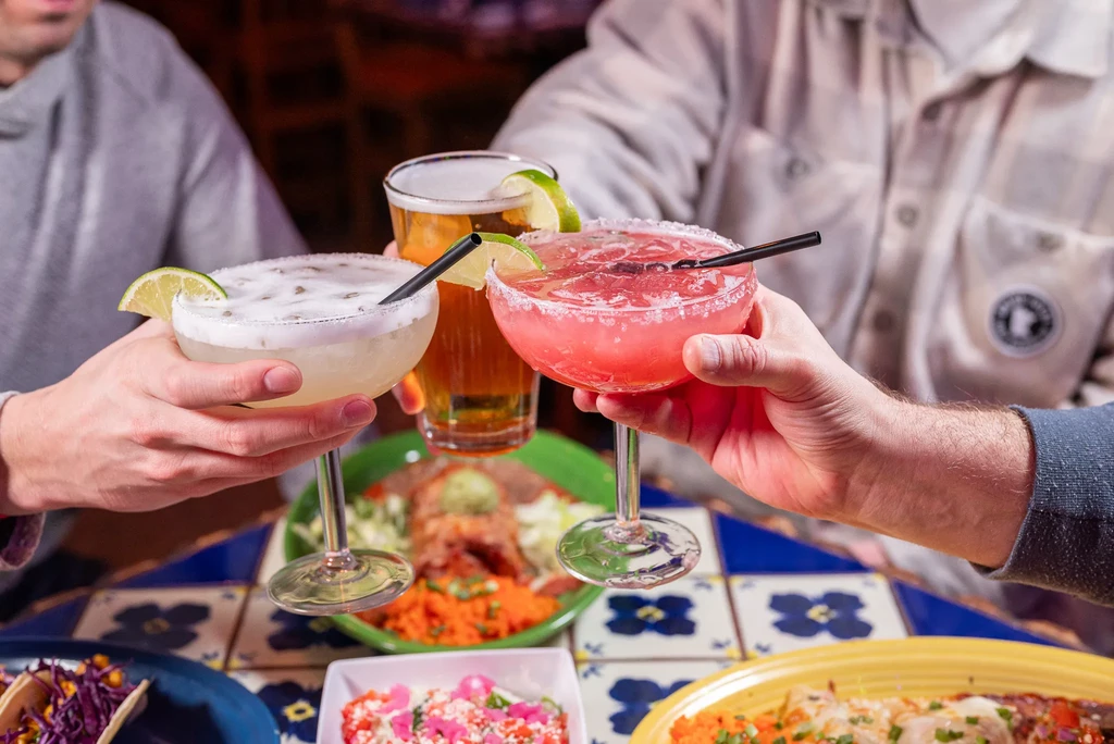 Three hands toast vibrant margaritas and beer above colorful food.