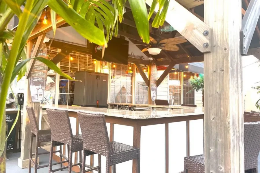 Tropical outdoor bar with wooden counter, stools, and palm fronds.