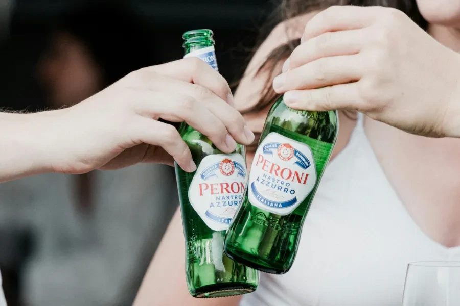 Two hands clinking green Peroni Nastro Azzurro beer bottles.