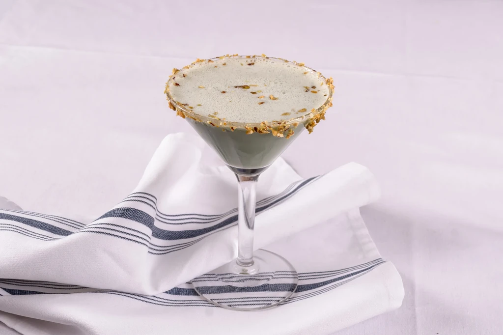 Martini glass with frothy light green drink, nut-garnished rim, on striped napkin.