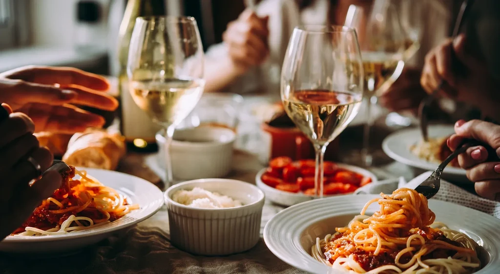 Warm close-up of hands, spaghetti, and wine on a dining table.