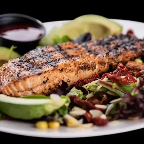 Grilled salmon with avocado, salad greens, and a side of sauce.