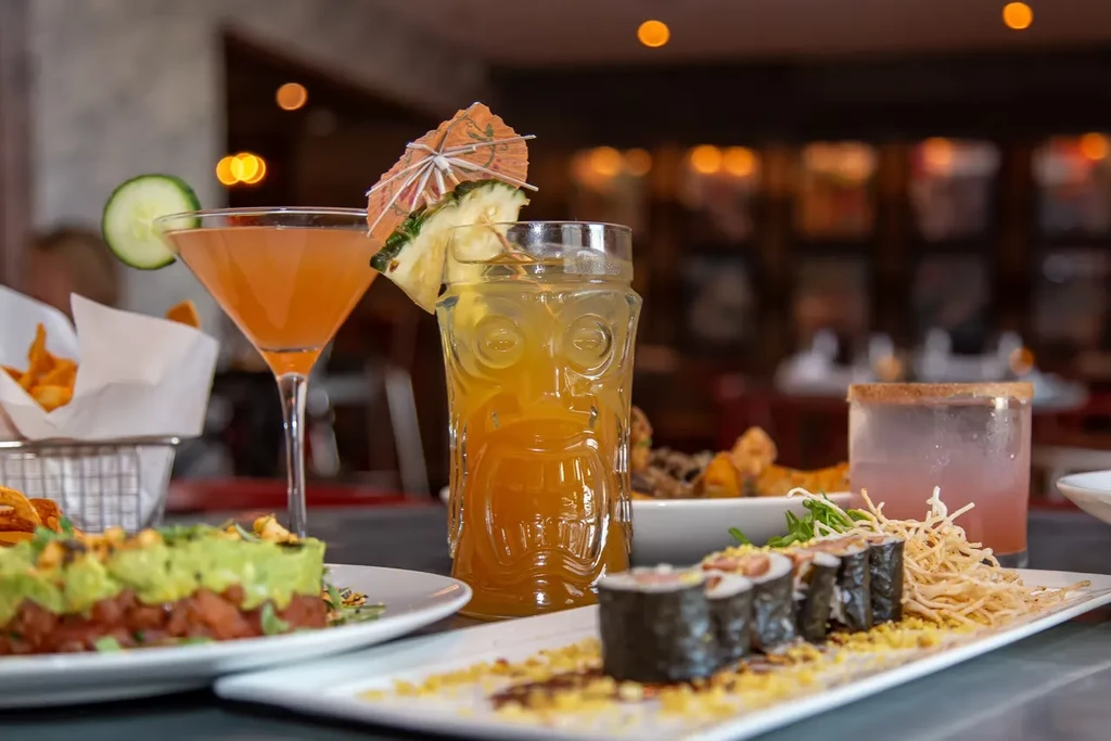 Amber martini, tiki drink, sushi, and layered dish on restaurant table.