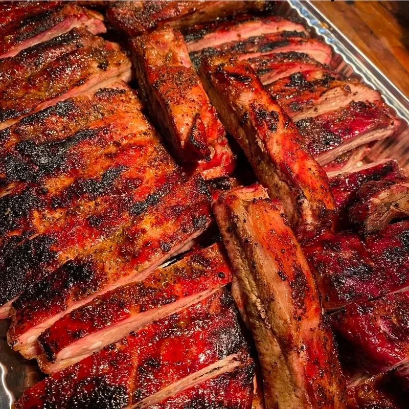 Numerous glistening, reddish-brown barbecue ribs on a metal tray.