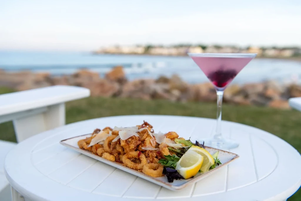 Fried calamari and pink cocktail on an Adirondack table by water.