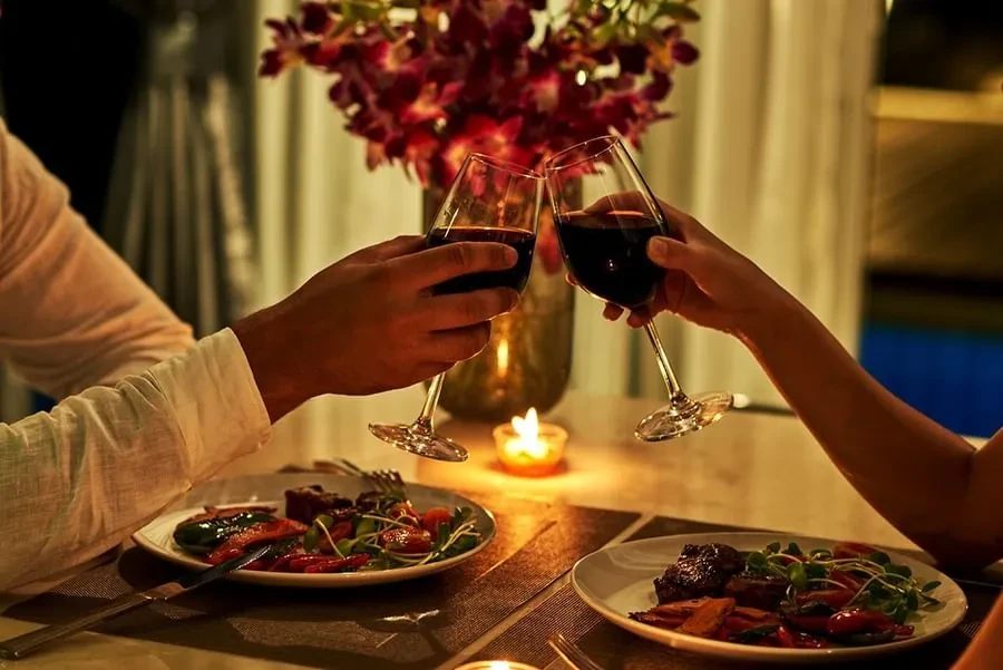 Hands clinking red wine glasses over a candlelit dinner table.