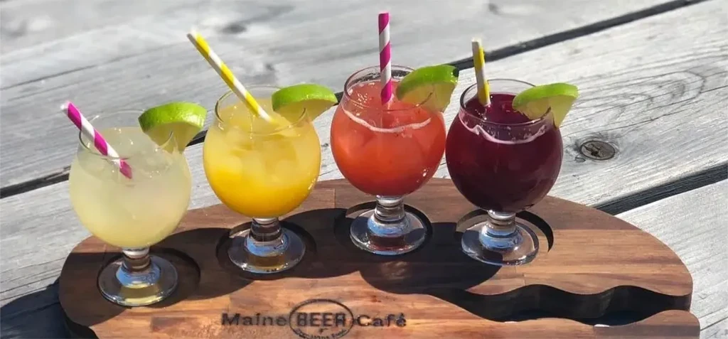 Four colorful drinks with lime, straws, on 'Maine BEER Café' tray.