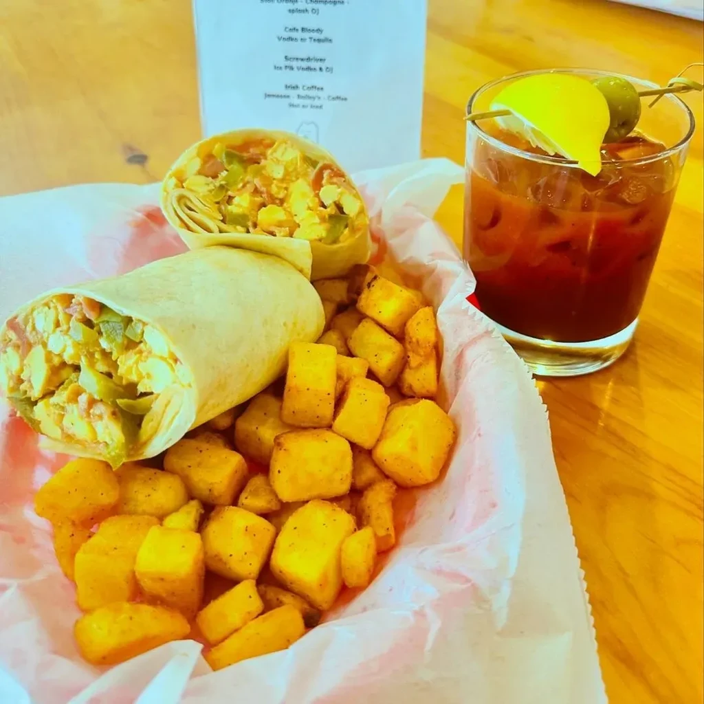 Breakfast burritos, cubed potatoes, and a garnished Bloody Mary on a wooden table.