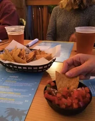 Hand dipping tortilla chip into salsa, with beer and a menu.