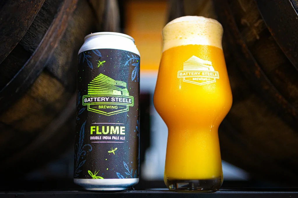 Battery Steele Brewing Flume IPA can and glass with blurred barrels.
