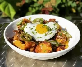 White bowl of roasted potatoes, sunny-side up egg, chives, and jalapeños.