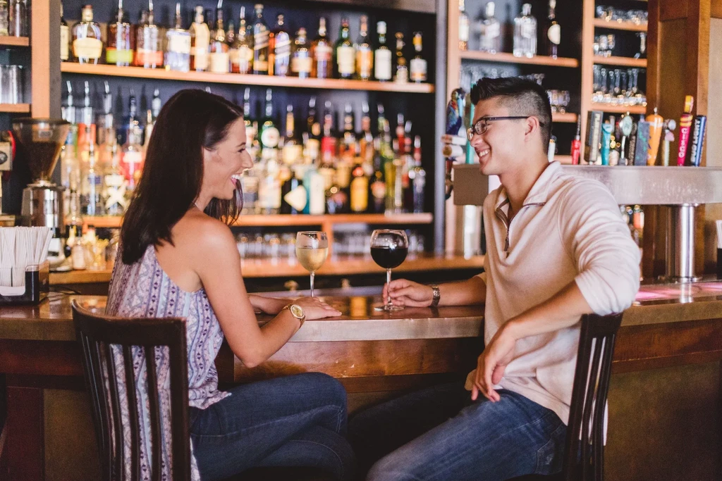 Man and woman smiling, drinking wine at a well-stocked bar.
