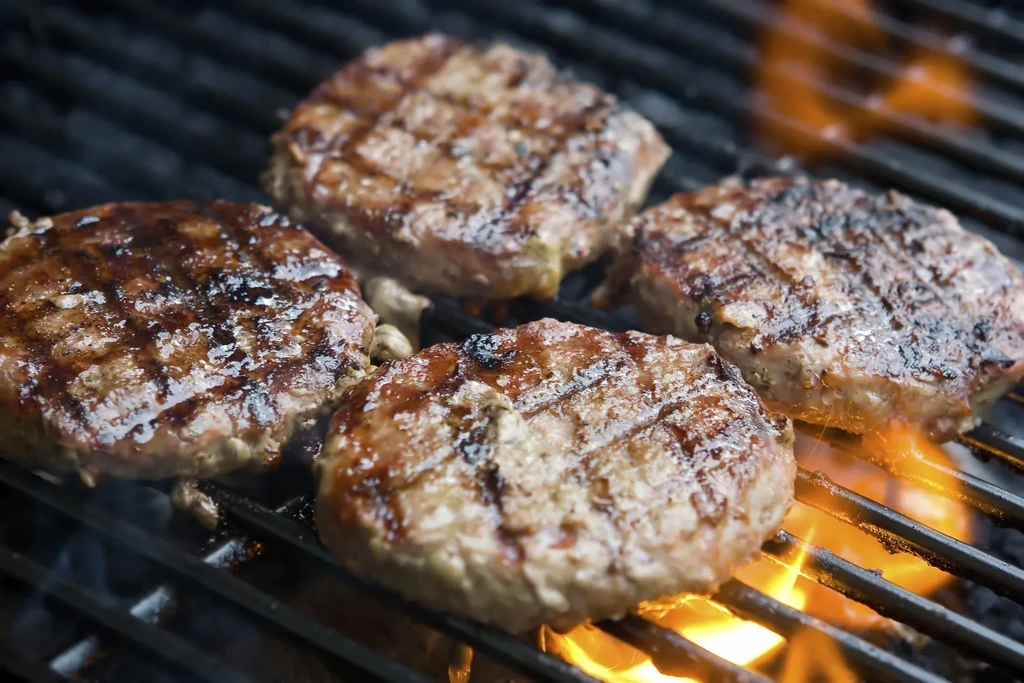 Four meat patties with grill marks cooking over open flames.