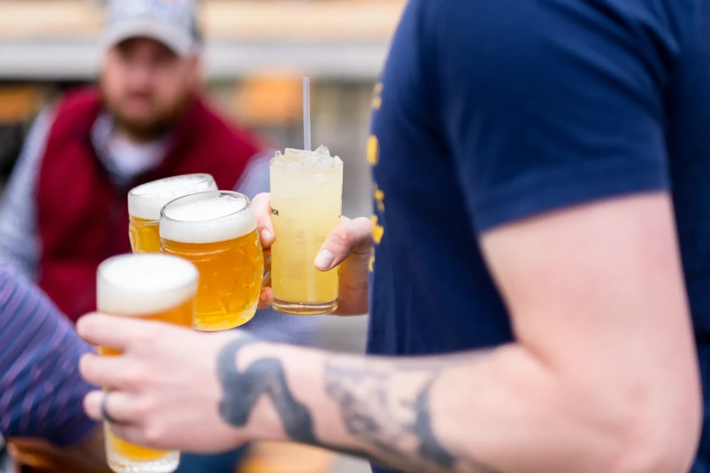 Hands holding three frothy beers and a yellow cocktail, one tattooed.