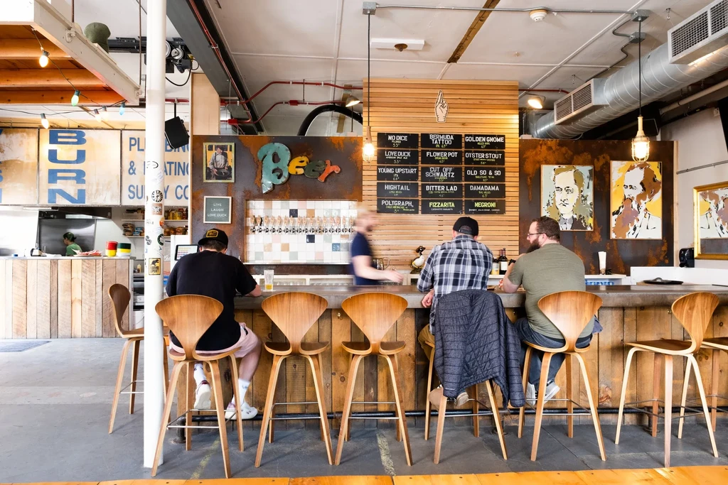 Bustling industrial bar interior with patrons and a bartender.