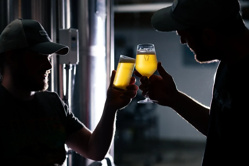 Two silhouetted people clink beer glasses in a backlit brewery.