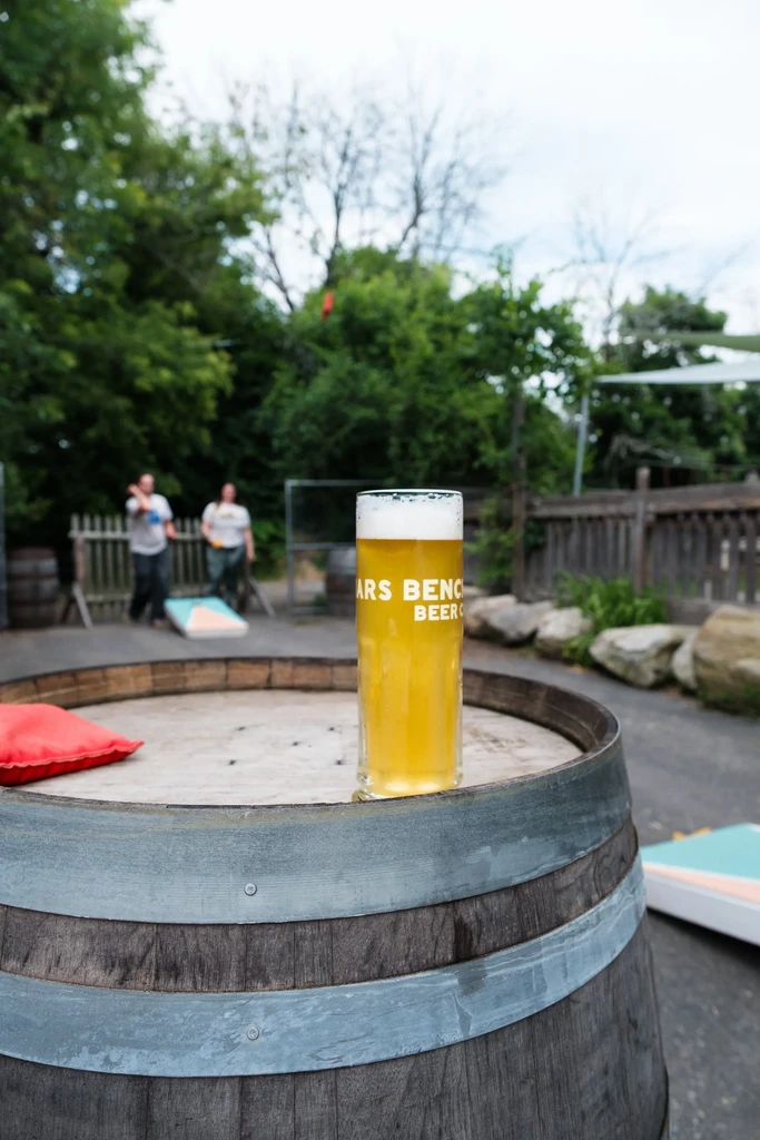 ARS BENCH BEER CO glass on barrel; people play cornhole outdoors.
