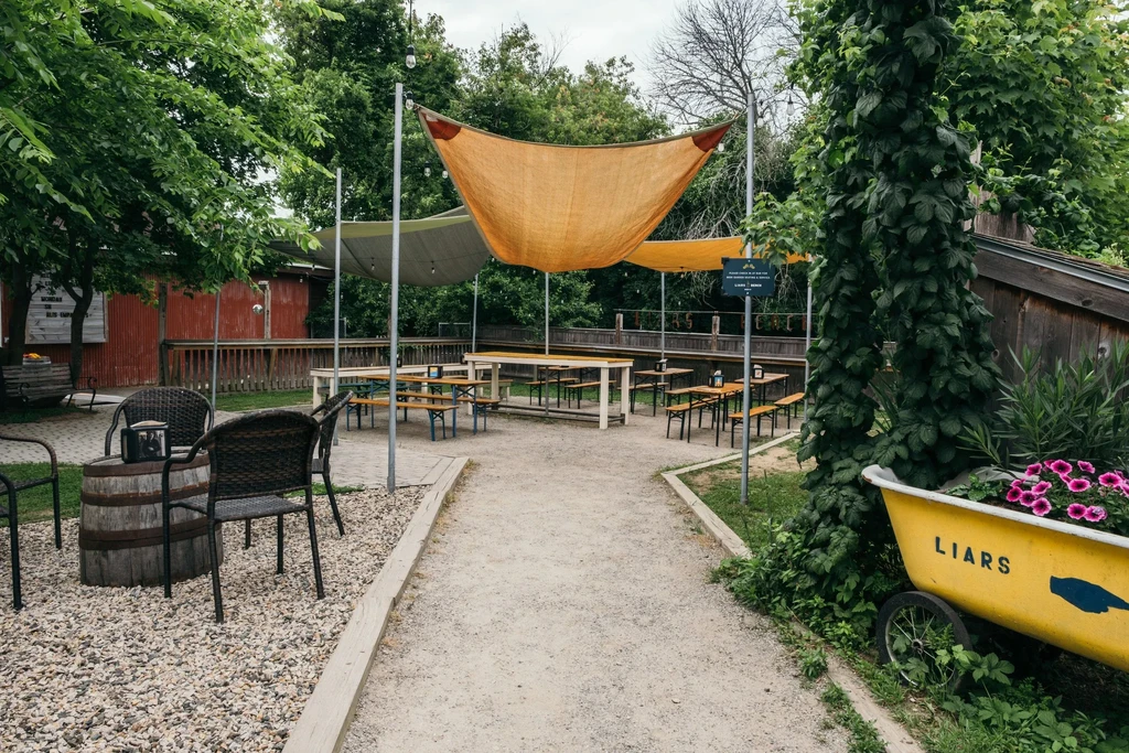 Rustic outdoor dining with picnic tables, shade sails, and a "LIARS" planter.