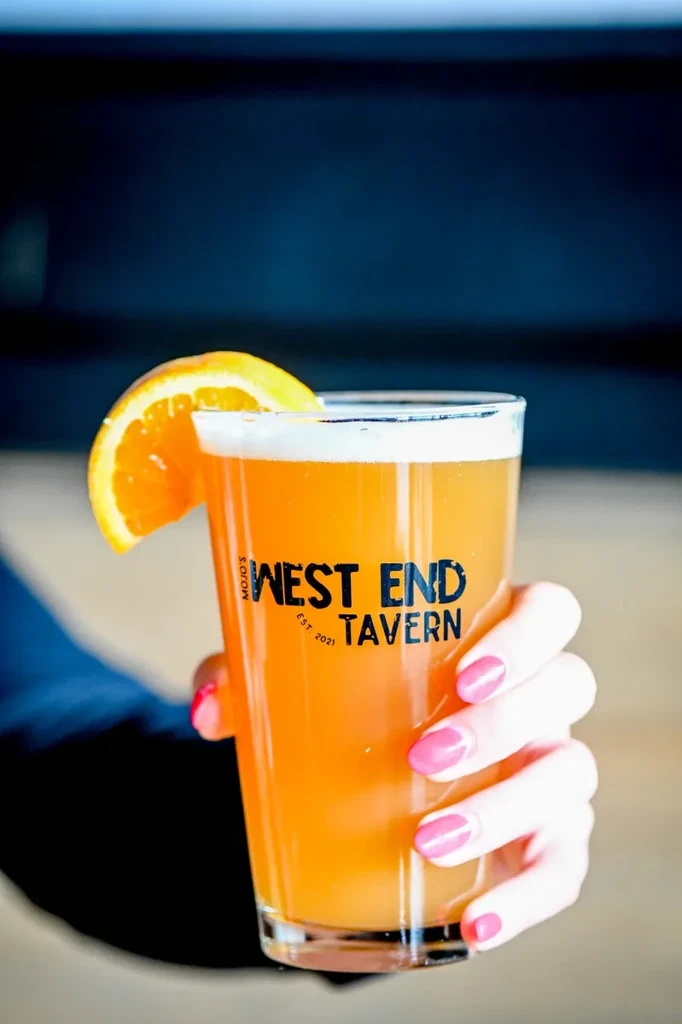 Hand holds West End Tavern glass with amber drink and orange slice.