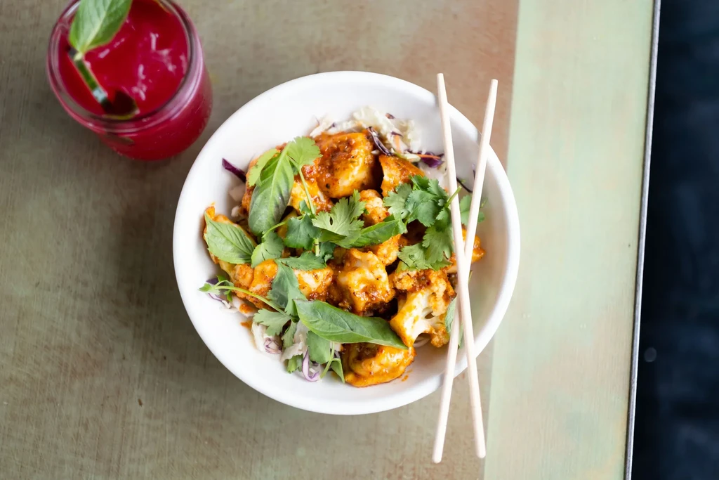 Overhead of roasted cauliflower with herbs, chopsticks, and a red drink.