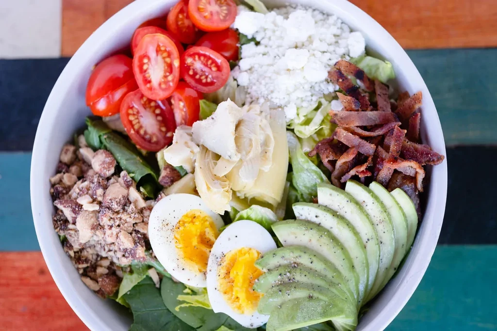 Overhead colorful salad with eggs, avocado, bacon, feta, and tomatoes.