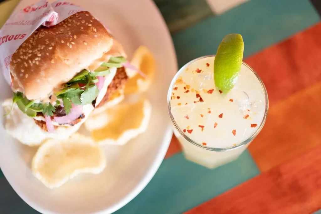 Sandwich, crispy items, avocado, and garnished cocktail on a striped table.