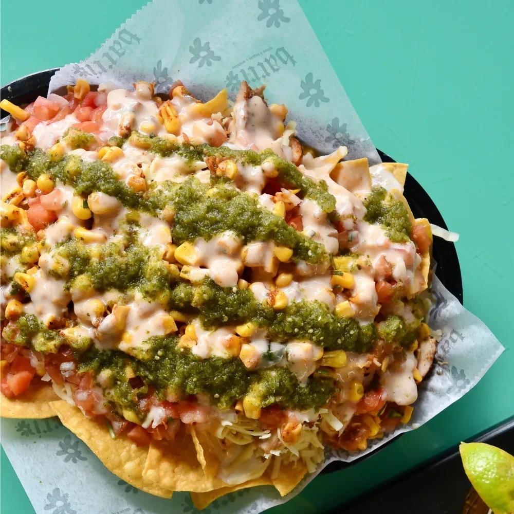 Nachos in black bowl with cheese, corn, tomatoes, green salsa.
