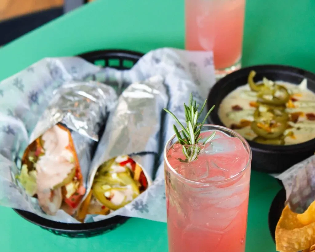 Drinks, foil-wrapped burritos, and jalapeño queso on a teal table.