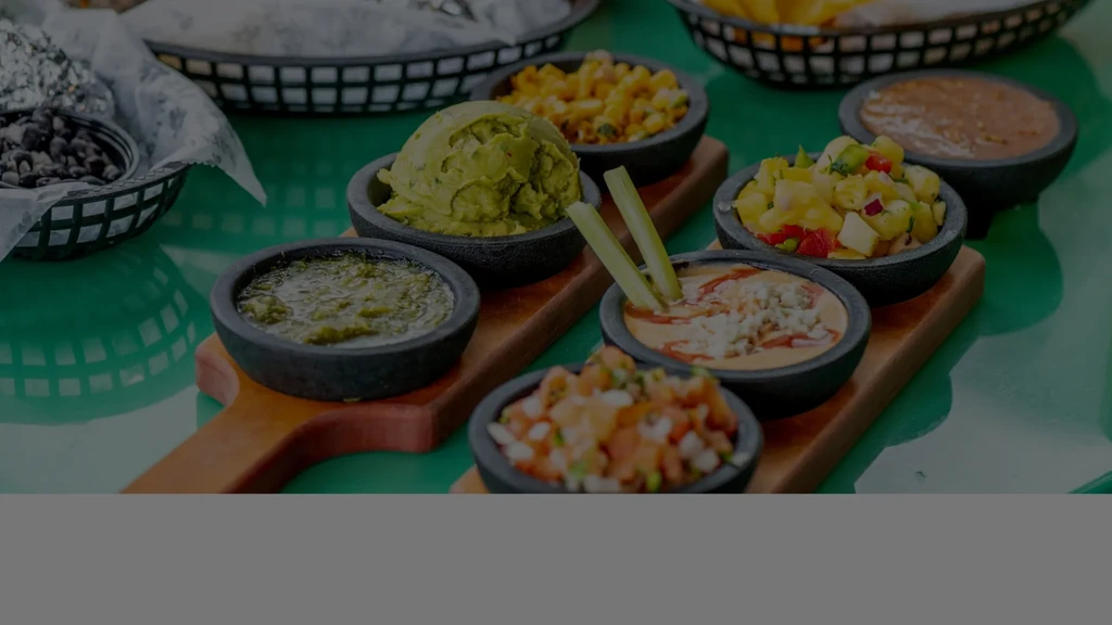 Seven bowls of colorful dips and salsas on a green table.