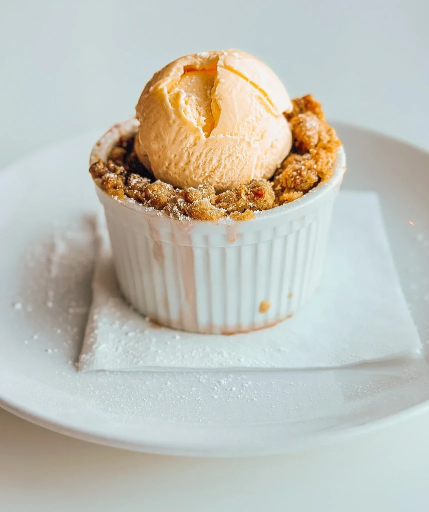 Apple crisp in ramekin with peach ice cream and powdered sugar.
