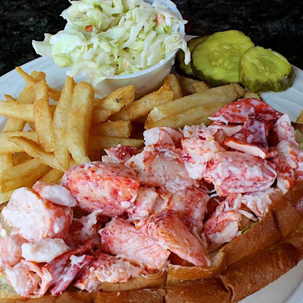 Lobster roll, french fries, coleslaw, and pickles served on a white plate.