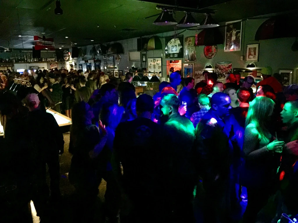 Crowded bar interior with colorful spotlights, a pool table, and sports decor.