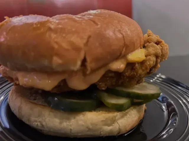 Close-up: golden fried chicken sandwich, creamy orange sauce, pickles.