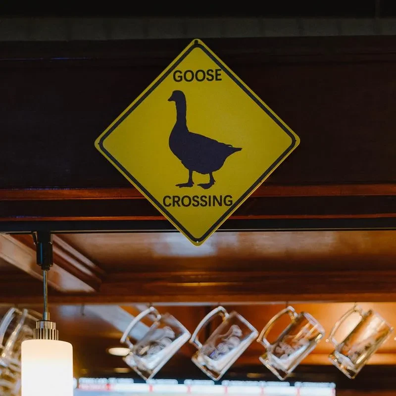 Yellow diamond 'GOOSE CROSSING' sign above mugs in a bar.