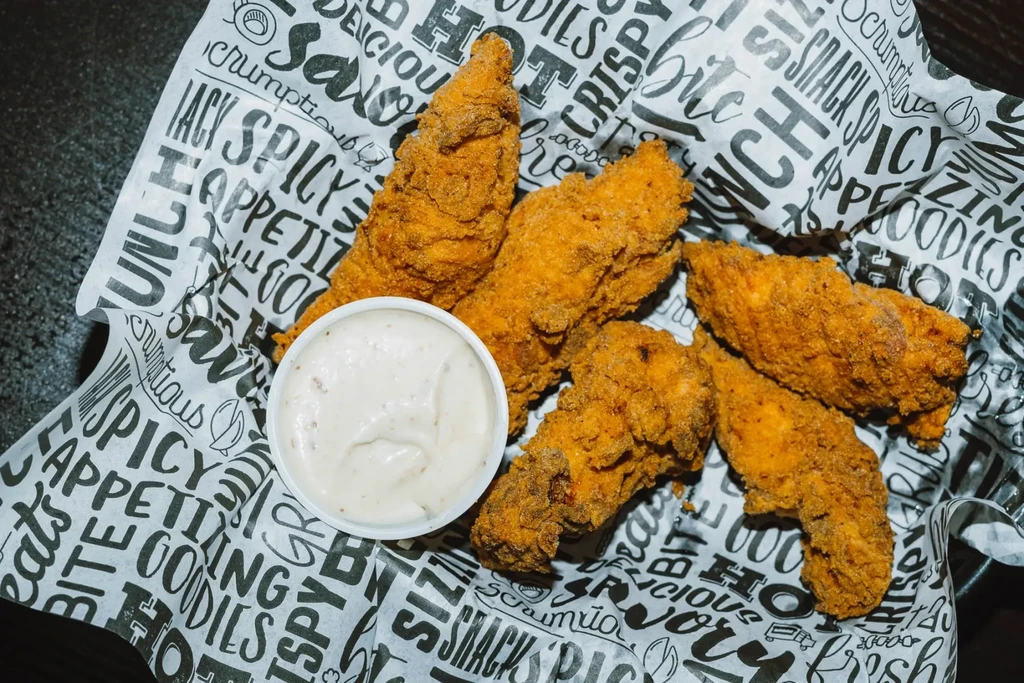 Crispy fried chicken tenders and creamy dipping sauce on word-printed paper.