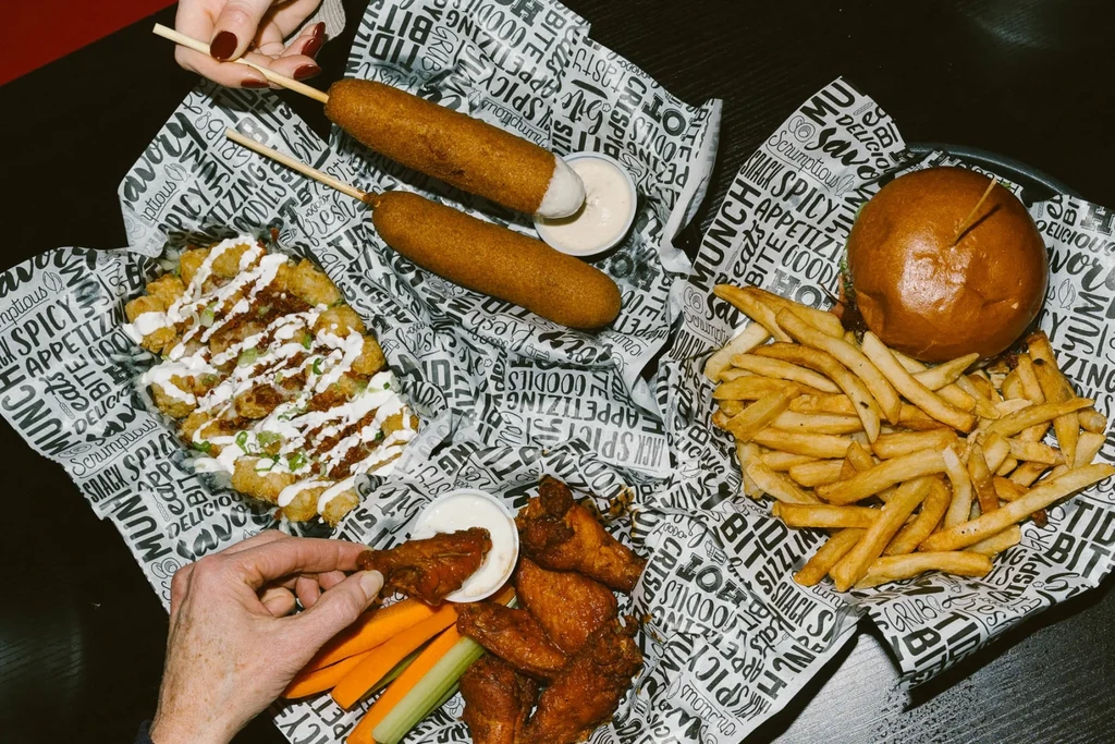 Overhead view: Hands eating baskets of corn dogs, fries, burger, and wings.
