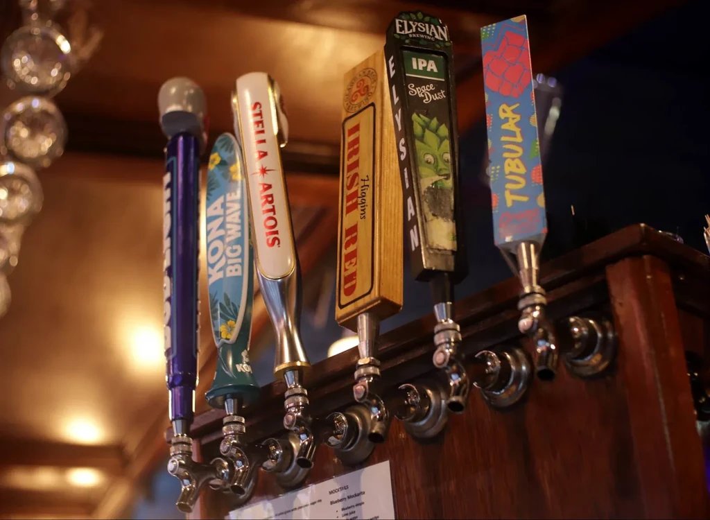 Six branded beer tap handles on a wooden bar counter.