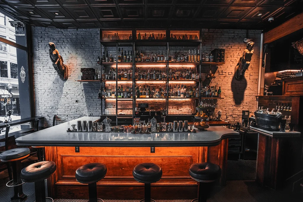 Dimly lit bar interior with liquor shelves, bartending tools, and carved figures.