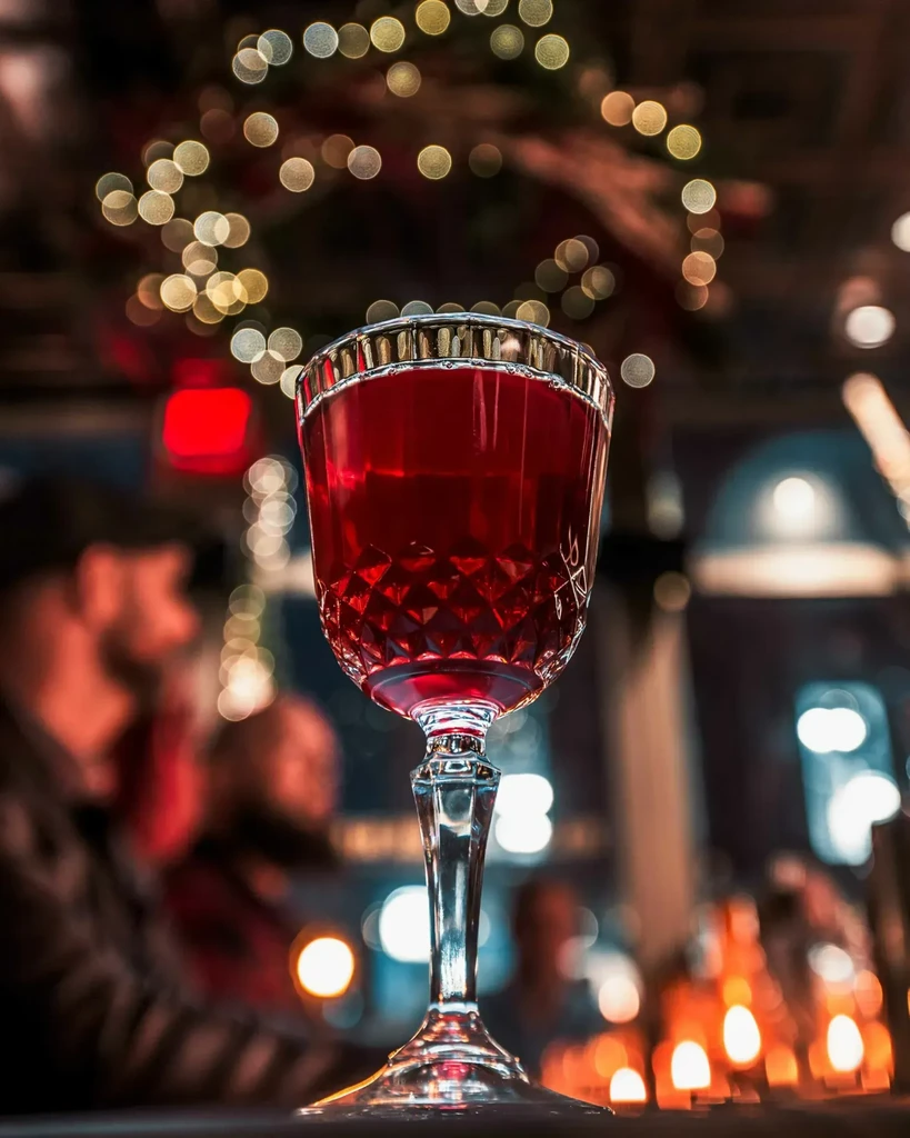 Faceted glass with red drink, blurry warm lights and figures.