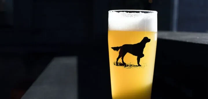 Amber beer in glass with frothy head and black dog silhouette.