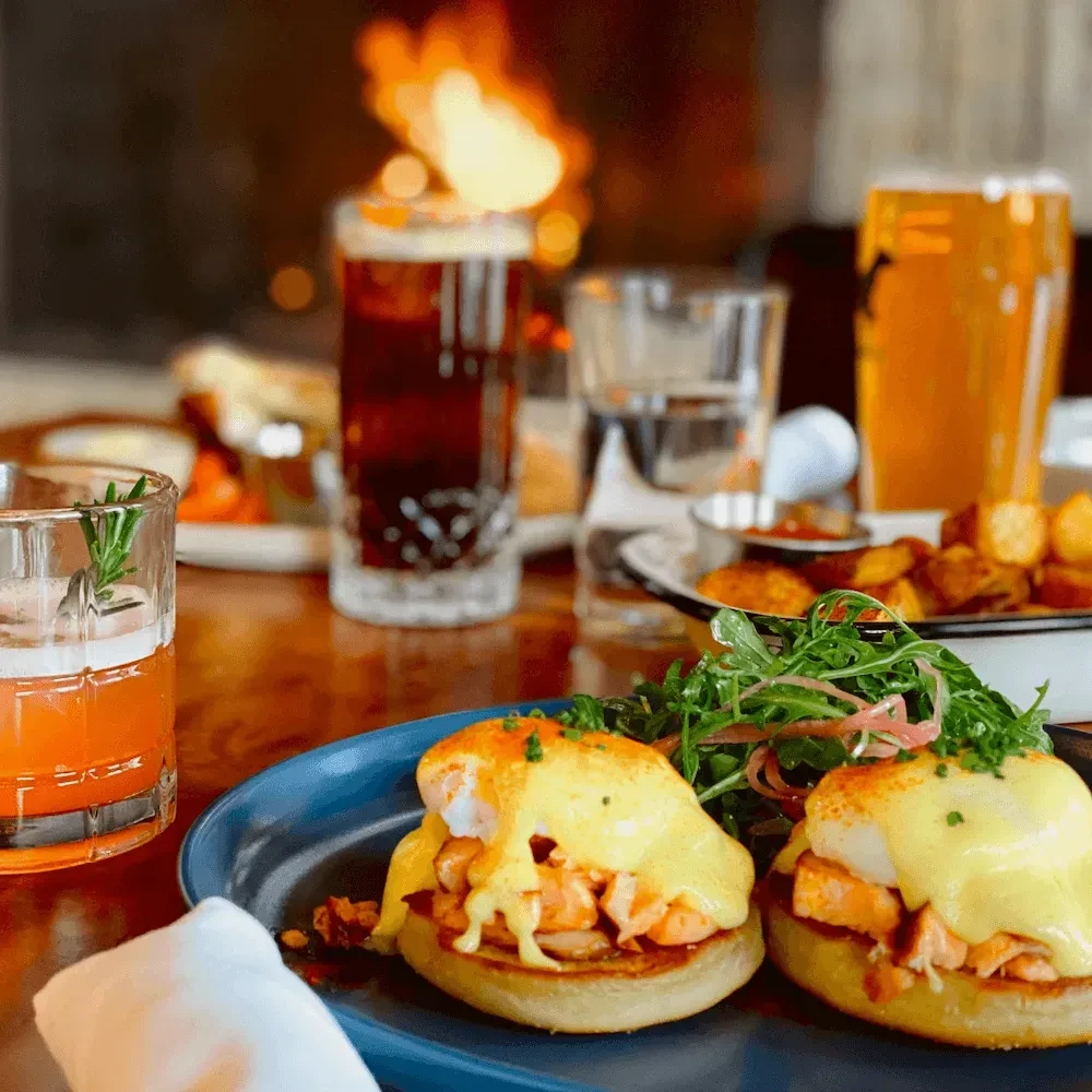Two Eggs Benedict, cocktail, and a blurred background fireplace.